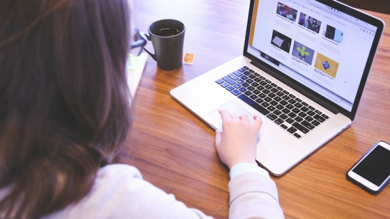 Eine Person mit langen Haaren und Brille benutzt einen Laptop auf einem Holztisch. Der Laptop-Bildschirm, möglicherweise gefüllt mit Schlüsselwörtern für Pinterest, zeigt mehrere Bilder. Ein Smartphone und eine Kaffeetasse liegen ebenfalls auf dem Tisch, während die Person mit der rechten Hand das Touchpad bedient.