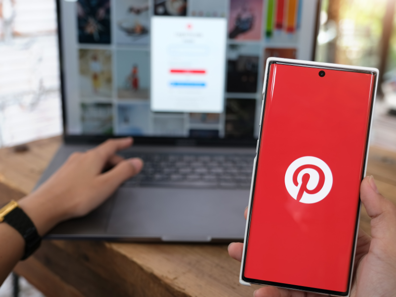 Eine Person hält ein Smartphone mit dem Pinterest-Logo vor einen Laptop. Dieser zeigt einen verschwommenen Pinterest-Anmeldebildschirm und ein Fotoraster. Die Szene auf einem Holztisch im Innenbereich zeigt eine ideale Pin-Beschreibung.