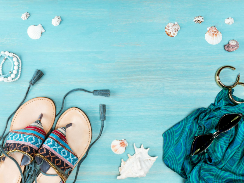Sommerlich inspiriertes Flatlay mit Strandthema auf blauem Holzhintergrund mit bunten Sandalen, gemustertem blaugrünem Stoff, Sonnenbrille, Armband und Muscheln.