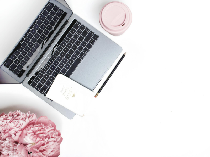 Auf einer weißen Oberfläche steht ein aufgeklappter Laptop, daneben ein schwarzer Bleistift, ein „Busy“-Notizbuch und ein rosafarbener Mehrwegbecher. Rosafarbene Blumen zieren die Ecke, während auf dem Bildschirm Texte angezeigt werden – das perfekte Tool für Ihren ordentlich organisierten und minimalistischen Arbeitsplatz.