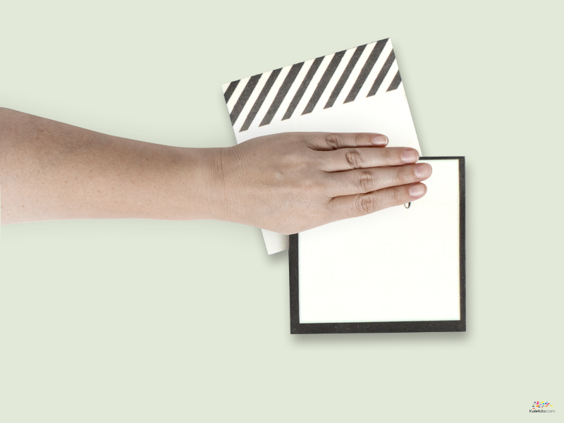 Eine Hand hält diskret zwei leere Karten: eine mit diagonalen schwarzen Streifen und die andere schlicht mit schwarzem Rand, der sich subtil vor einem hellgrünen Hintergrund versteckt.