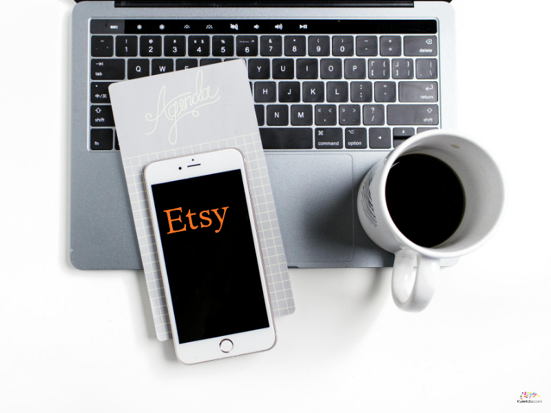 Auf der Tastatur ruht ein Laptop mit einem Notizblock und einem Smartphone, auf dem das Etsy-Logo deutlich zu sehen ist. Daneben trägt eine weiße Tasse mit schwarzem Kaffee zum gemütlichen Charme des Arbeitsbereichs bei.