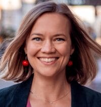 Eine Frau mit schulterlangem hellbraunem Haar lächelt draußen in einer urbanen Umgebung. Sie trägt rote Perlenohrringe, einen dunklen Blazer und ein rosa Top – ihr selbstbewusster Look ist perfekt für ein Pin-Titel-Profilfoto.