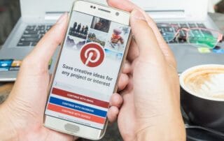 Eine Person hält ein Smartphone mit dem Pinterest-Anmeldebildschirm in der Hand und ist bereit für ein Pinterest-Audit. Sie hat die Wahl, per E-Mail, Facebook oder Google fortzufahren. Im unscharfen Hintergrund vervollständigen ein Laptop und eine dampfende Tasse Latte die Szene.