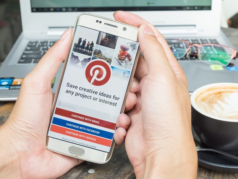Eine Person hält ein Smartphone mit dem Pinterest-Anmeldebildschirm in der Hand und ist bereit für ein Pinterest-Audit. Sie hat die Wahl, per E-Mail, Facebook oder Google fortzufahren. Im unscharfen Hintergrund vervollständigen ein Laptop und eine dampfende Tasse Latte die Szene.