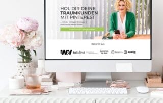 Ein Computerbildschirm auf einem weißen Schreibtisch zeigt eine deutschsprachige Webseite für Pinterest-Marketing. Eine Frau in einem grünen Blazer lächelt, darunter befinden sich Logos. Auf dem Schreibtisch stehen eine Vase mit rosa Blumen und verschiedene Büroartikel, die der Szenerie Charme verleihen.