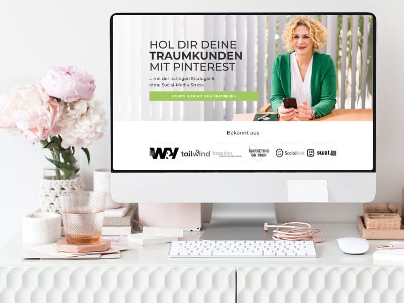 Ein Computerbildschirm auf einem weißen Schreibtisch zeigt eine deutschsprachige Webseite für Pinterest-Marketing. Eine Frau in einem grünen Blazer lächelt, darunter befinden sich Logos. Auf dem Schreibtisch stehen eine Vase mit rosa Blumen und verschiedene Büroartikel, die der Szenerie Charme verleihen.