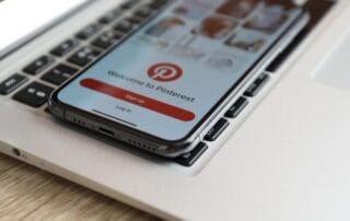 Ein Smartphone mit dem Pinterest-Anmeldebildschirm liegt auf der Tastatur eines silbernen Laptops und zeigt, wie sich Pinterest nahtlos in Ihren Arbeitsablauf integrieren lässt. Auf dem Telefonbildschirm werden Optionen zur Anmeldung oder Anmeldung bei Pinterest angezeigt.