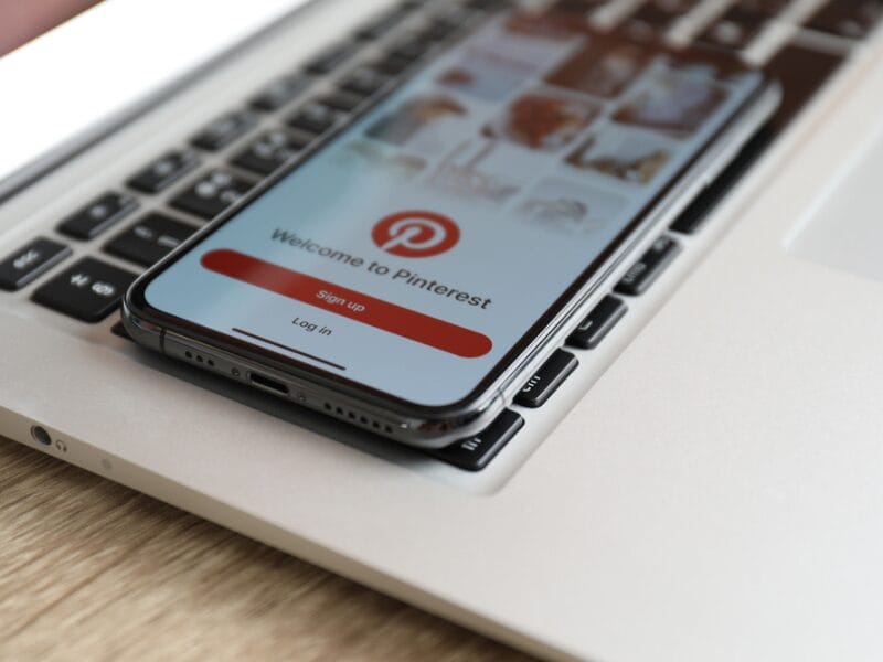 Ein Smartphone mit dem Pinterest-Anmeldebildschirm liegt auf der Tastatur eines silbernen Laptops und zeigt, wie sich Pinterest nahtlos in Ihren Arbeitsablauf integrieren lässt. Auf dem Telefonbildschirm werden Optionen zur Anmeldung oder Anmeldung bei Pinterest angezeigt.
