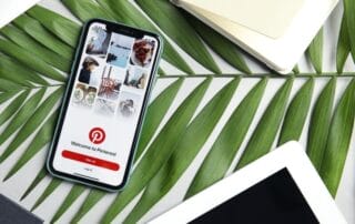 Ein Smartphone mit dem Pinterest-Anmeldebildschirm ruht auf grünen Palmblättern, umgeben von einem weißen Tablet, einem geschlossenen Notizbuch und einem schwarzen Objekt – die perfekte Inspiration für Ihren nächsten Blogartikel zum Thema stilvolle Arbeitsplätze.