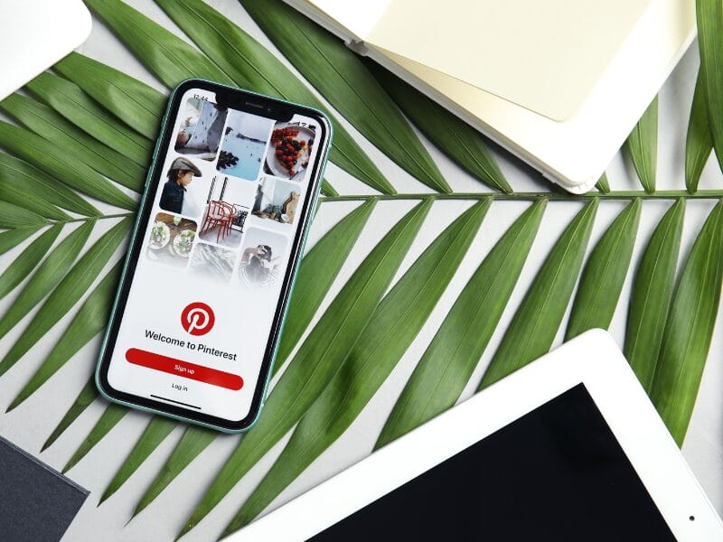 Ein Smartphone mit dem Pinterest-Anmeldebildschirm ruht auf grünen Palmblättern, umgeben von einem weißen Tablet, einem geschlossenen Notizbuch und einem schwarzen Objekt – die perfekte Inspiration für Ihren nächsten Blogartikel zum Thema stilvolle Arbeitsplätze.