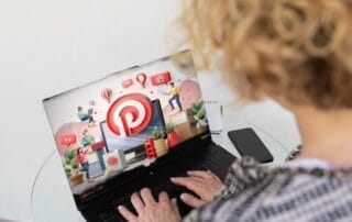 Eine Person mit lockigem blondem Haar benutzt einen Laptop, auf dem das Pinterest-Logo abgebildet ist, umgeben von digitalen Symbolen und Grafiken. Der Laptop steht auf einem Glastisch, in dessen Nähe sich ein Smartphone befindet, was auf die Konsequenz hinweist, in einer digitalen Welt in Verbindung zu bleiben.