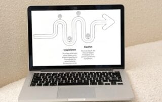 Ein Laptop auf einer beigen Oberfläche zeigt einen Bildschirm mit einer schwarz-weißen Kundenreise-Grafik - ein verschlungener Pfad, der in einem Pfeil nach rechts endet, neben einem deutschen Text, der die Schritte zur Inspiration und zum Kauf erklärt.