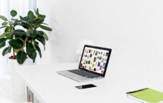 Ein aufgeräumter weißer Schreibtisch mit einem Laptop, auf dem ein Raster aus bunten nischen Buchdeckeln zu sehen ist, ein Smartphone, ein grünes Notizbuch mit einem Stift und eine blattreiche Topfpflanze im Hintergrund.