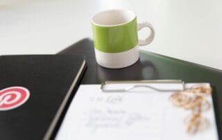 Ein grün-weißer Kaffeebecher steht auf einem geschlossenen Laptop neben einem schwarzen Notizbuch mit einem roten Aufkleber mit der Aufschrift "Löschen". Im Vordergrund ist ein Klemmbrett mit Papier und goldenen Büroklammern leicht unscharf.