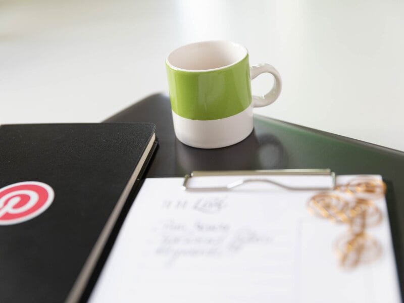 Ein grün-weißer Kaffeebecher steht auf einem geschlossenen Laptop neben einem schwarzen Notizbuch mit einem roten Aufkleber mit der Aufschrift "Löschen". Im Vordergrund ist ein Klemmbrett mit Papier und goldenen Büroklammern leicht unscharf.
