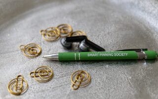 Ein grüner Stift mit der Aufschrift "SMART PINNING SOCIETY" liegt auf einer strukturierten silbernen Oberfläche, umgeben von goldenen Drahtspiralen und einem Paar schwarzer kabelloser Ohrhörer - perfekt, um deine Gründe stilvoll zu notieren.
