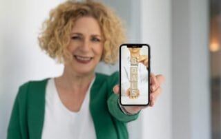 Eine lächelnde Frau mit lockigem blondem Haar hält ein Smartphone in die Kamera. Auf dem Bildschirm des Telefons sind vertikal gestapelte Holzklötze zu sehen, auf denen "GOALS" - oder "Ziele" - steht. Die Frau trägt eine grüne Strickjacke und ein weißes Hemd.