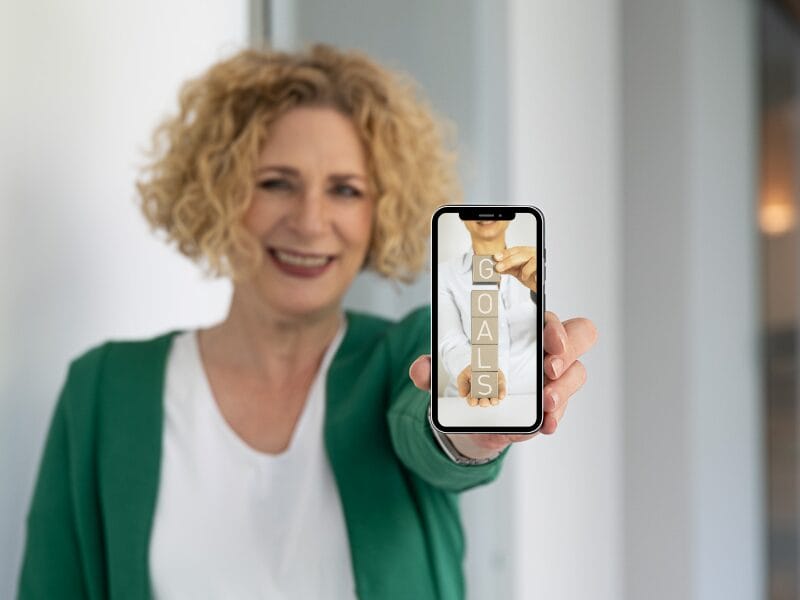 Eine lächelnde Frau mit lockigem blondem Haar hält ein Smartphone in die Kamera. Auf dem Bildschirm des Telefons sind vertikal gestapelte Holzklötze zu sehen, auf denen "GOALS" - oder "Ziele" - steht. Die Frau trägt eine grüne Strickjacke und ein weißes Hemd.