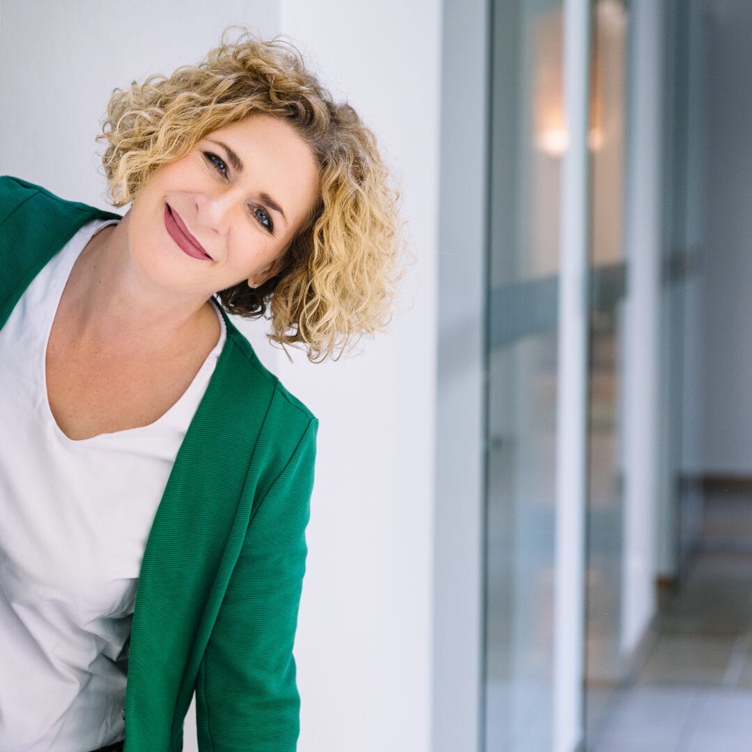 Barbara Riedl-Wiesinger Ueber uns Eine lächelnde Frau mit lockigem blondem Haar, die einen grünen Blazer und ein weißes Oberteil trägt, lehnt sich in einem hellen Flur mit Glaswänden leicht zur Seite und verkörpert den aufgeschlossenen und professionellen Geist einer Assistenz.