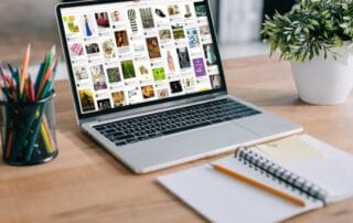 Auf einem hölzernen Schreibtisch steht ein Laptop mit einem Raster von Bildern, daneben eine kleine Pflanze, ein Becher mit Buntstiften und ein aufgeschlagenes Notizbuch mit einem Stift - perfekt für die Planung Ihrer Pin-Zeiten und kreativen Projekte.