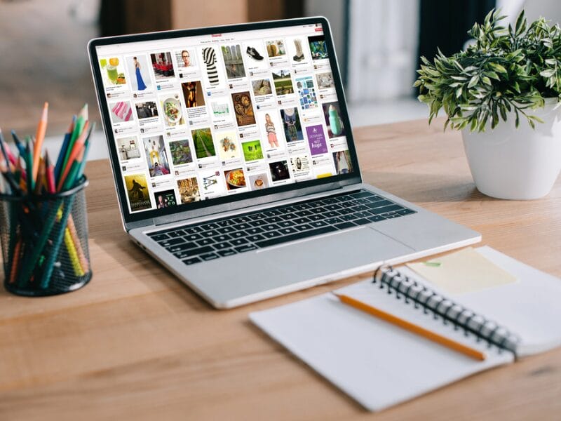 Auf einem hölzernen Schreibtisch steht ein Laptop mit einem Raster von Bildern, daneben eine kleine Pflanze, ein Becher mit Buntstiften und ein aufgeschlagenes Notizbuch mit einem Stift - perfekt für die Planung Ihrer Pin-Zeiten und kreativen Projekte.
