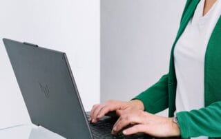 Eine Person, die einen grünen Blazer und ein weißes Hemd trägt, tippt auf einem schwarzen Laptop, der möglicherweise auf Pinterest-SEO recherchiert, auf einer Glasfläche vor einem schlichten weißen Hintergrund.
