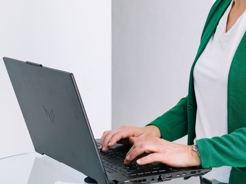 Eine Person, die einen grünen Blazer und ein weißes Hemd trägt, tippt auf einem schwarzen Laptop, der möglicherweise auf Pinterest-SEO recherchiert, auf einer Glasfläche vor einem schlichten weißen Hintergrund.