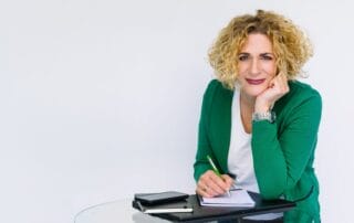 Eine Frau mit lockigem blondem Haar und grünem Blazer sitzt an einem Glastisch, lächelt und stützt ihren Kopf auf ihre Hand. Sie hält einen grünen Stift in der Hand und schreibt in ein Notizbuch, auf dem Tisch liegen ein Tablet und ein Telefon.