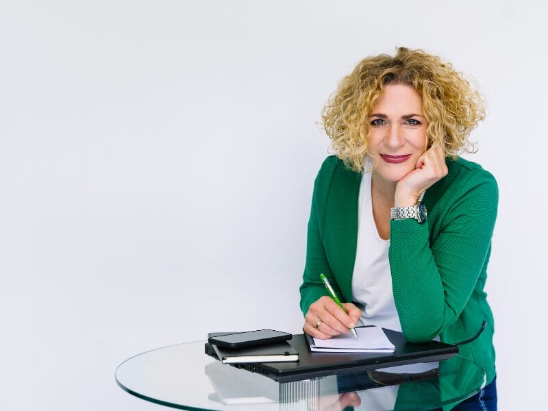 Eine Frau mit lockigem blondem Haar und grünem Blazer sitzt an einem Glastisch, lächelt und stützt ihren Kopf auf ihre Hand. Sie hält einen grünen Stift in der Hand und schreibt in ein Notizbuch, auf dem Tisch liegen ein Tablet und ein Telefon.