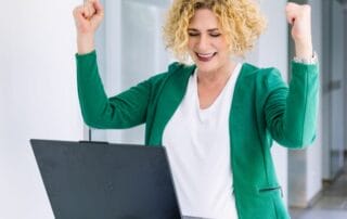 Eine Frau mit lockigen blonden Haaren, die einen grünen Blazer und ein weißes Hemd trägt, lächelt und hebt aufgeregt die Fäuste, während sie vor einem aufgeklappten Laptop steht - und einen Durchbruch in ihrer Wachstumsstrategie feiert.