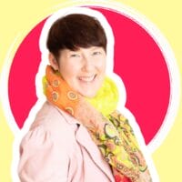 Eine lächelnde Frau mit kurzen dunklen Haaren, die eine hellrosa Jacke und einen bunt gemusterten Schal trägt, steht vor einem roten und gelben kreisförmigen Hintergrund - die perfekte Inspiration für Pinterest für Coaches, die lebendige, ansprechende Bilder suchen.