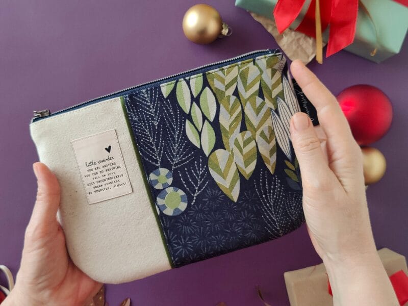 Gute Produktfotos für Pinterest gestalten 4 Eine Person hält einen Stoffbeutel mit Reißverschluss mit einem dekorativen Blatt- und geometrischen Muster, umgeben von Weihnachtsschmuck und Geschenkboxen auf einer lila Fläche - perfekt für festliche Produktfotos.