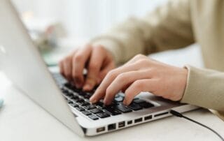 Nahaufnahme eines Anhängers, der an einem Schreibtisch auf einer Laptoptastatur tippt, wobei die Hände im Mittelpunkt stehen und der Bildschirm teilweise sichtbar ist. Die Person trägt ein helles, langärmeliges Oberteil.