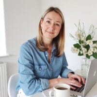 Eine Frau mit blonden Haaren und einem blauen Jeanshemd lächelt, während sie an einem weißen Tisch mit einer Tasse Kaffee und einer Vase mit weißen Blumen neben sich an einem Laptop arbeitet.